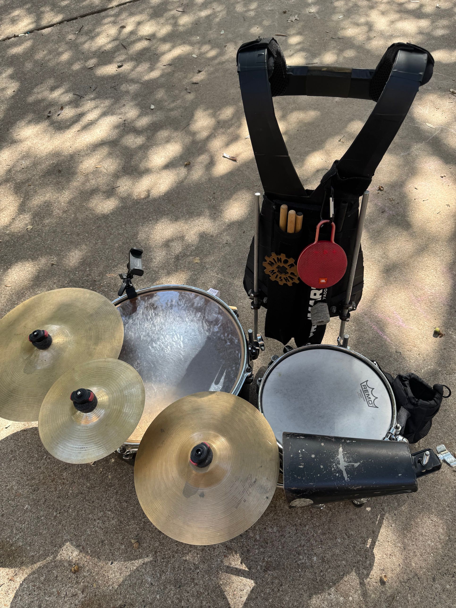 The assembled kit on a sidewalk, seen from the front and above: the marching harness stands behind two drums, with cymbals, a cowbell, a stick bag, a Minor Mishap badge and a clamp phone mount attached.