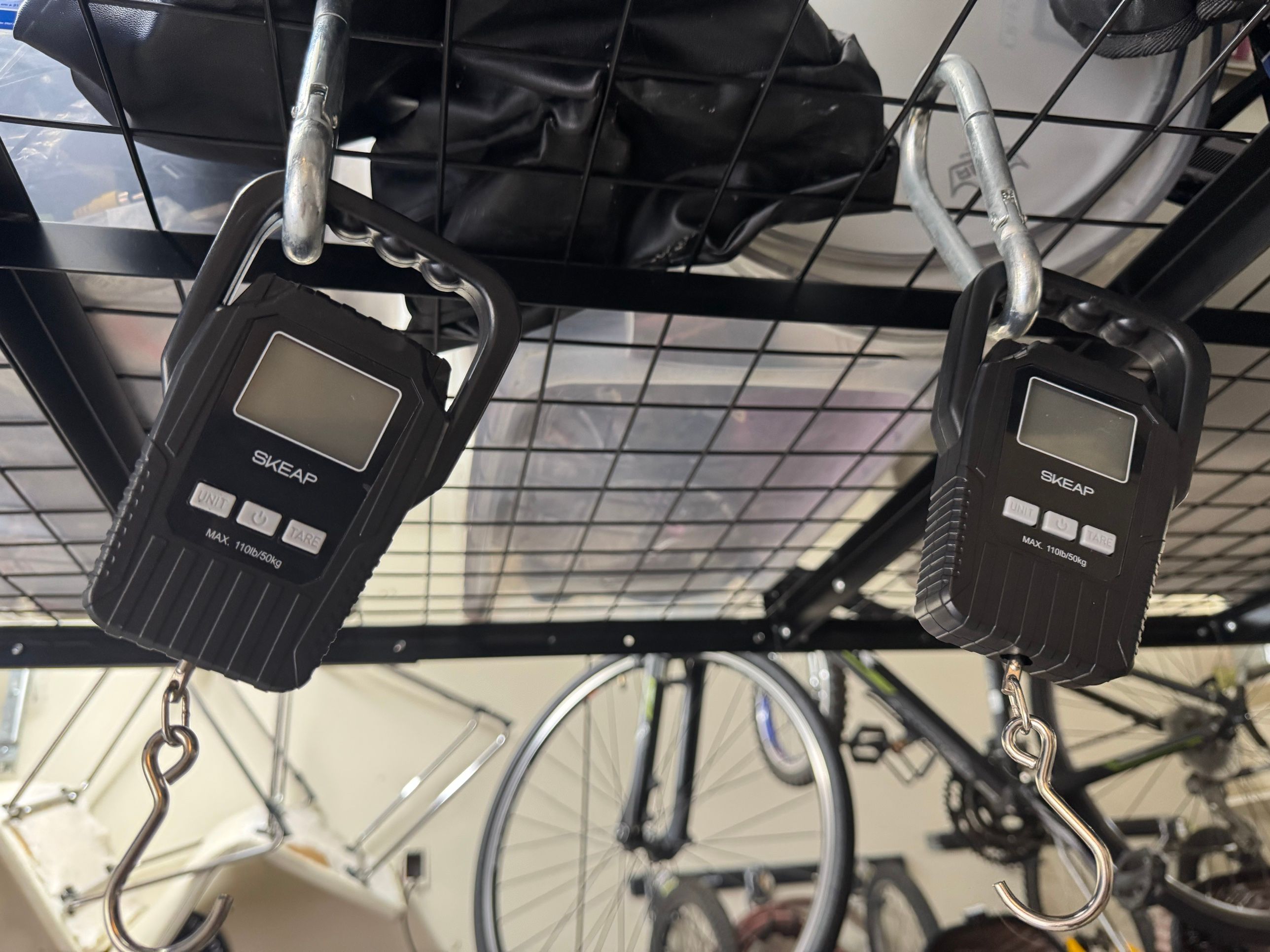 Two black SKEAP digital luggage scales hanging side by side from a ceiling rack, each marked "MAX. 110lb/50kg".